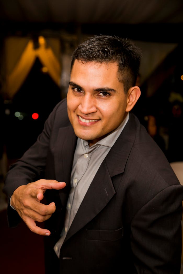 Portrait of a confident, smiling businessman in a formal suit pointing directly at the camera indoors.